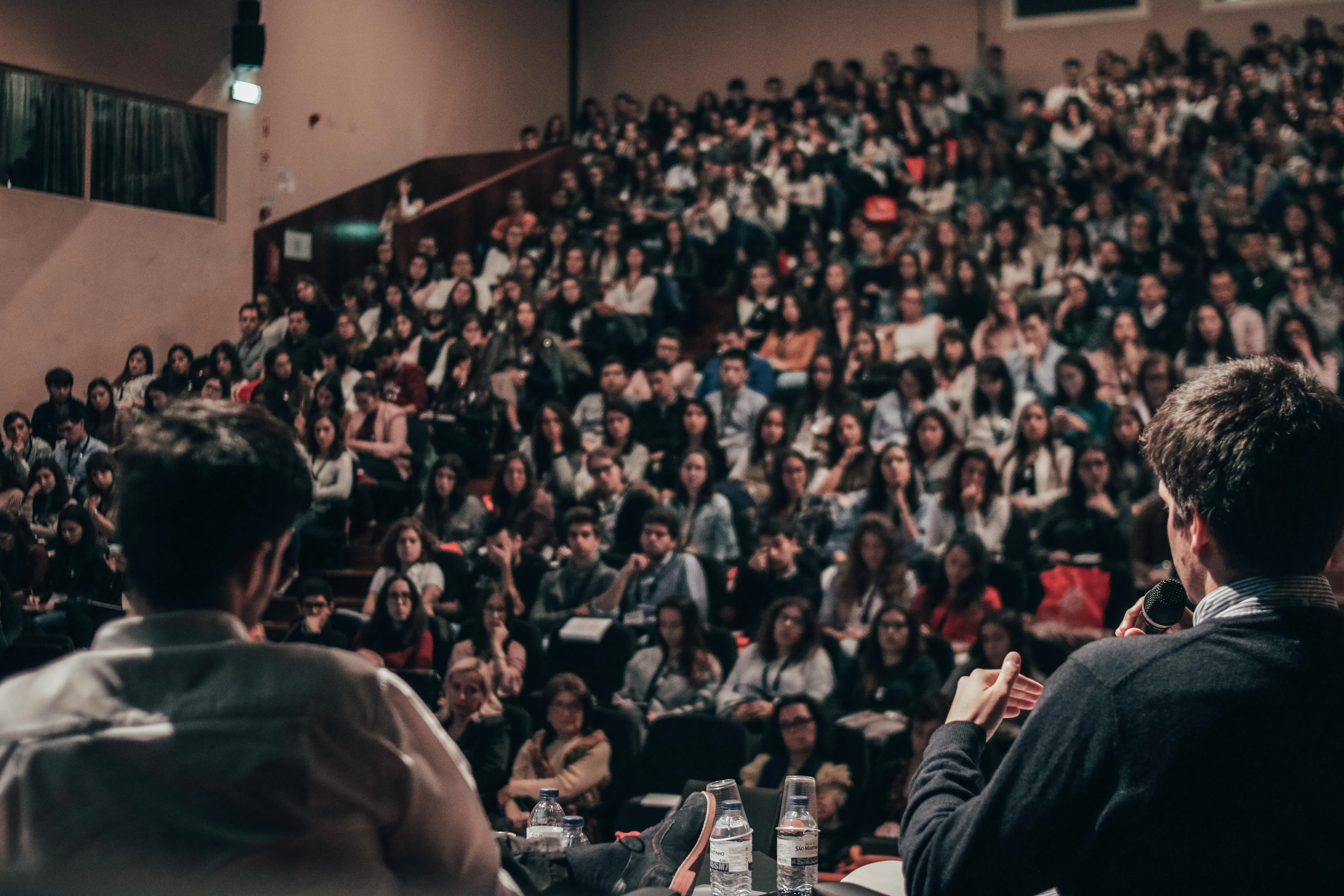 Uma plateia completa sentada em uma grande sala de aula, com dois palestrantes em primeiro plano