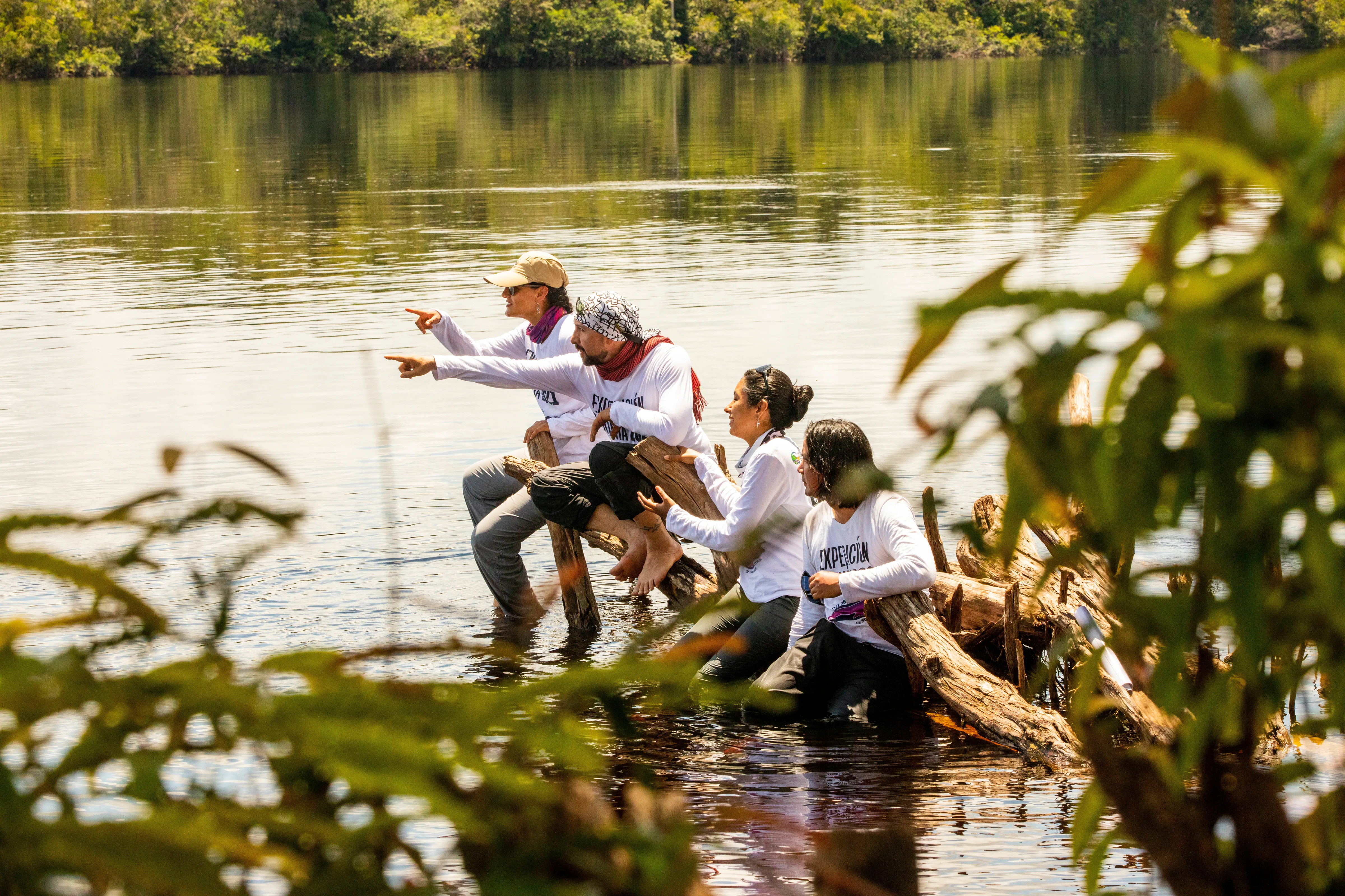 A group of people sitting on a log at the edge of a river, pointing into the distance