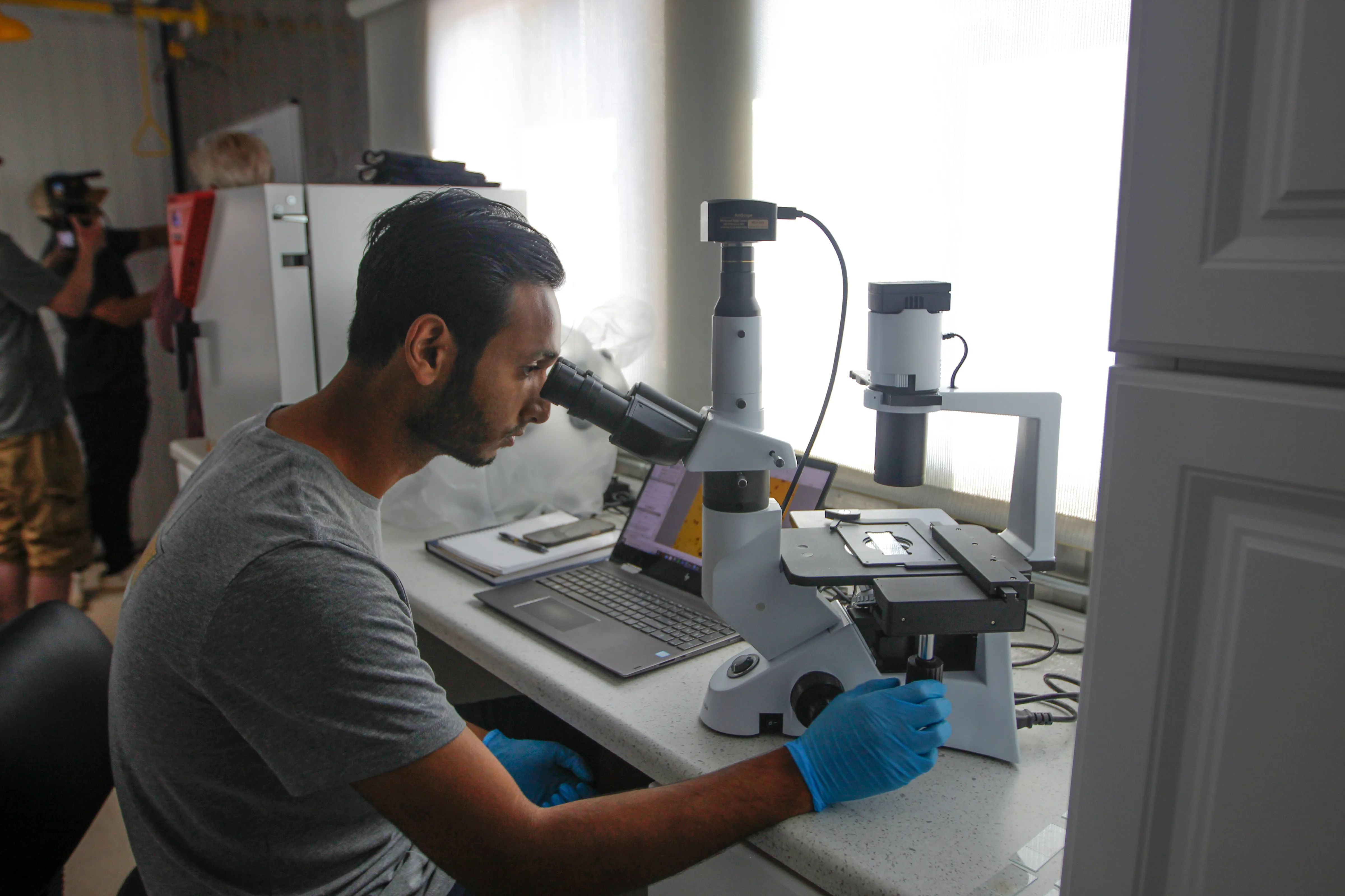 Um homem usando luvas de látex azuis em um laboratório branco, olhando através de um microscópio