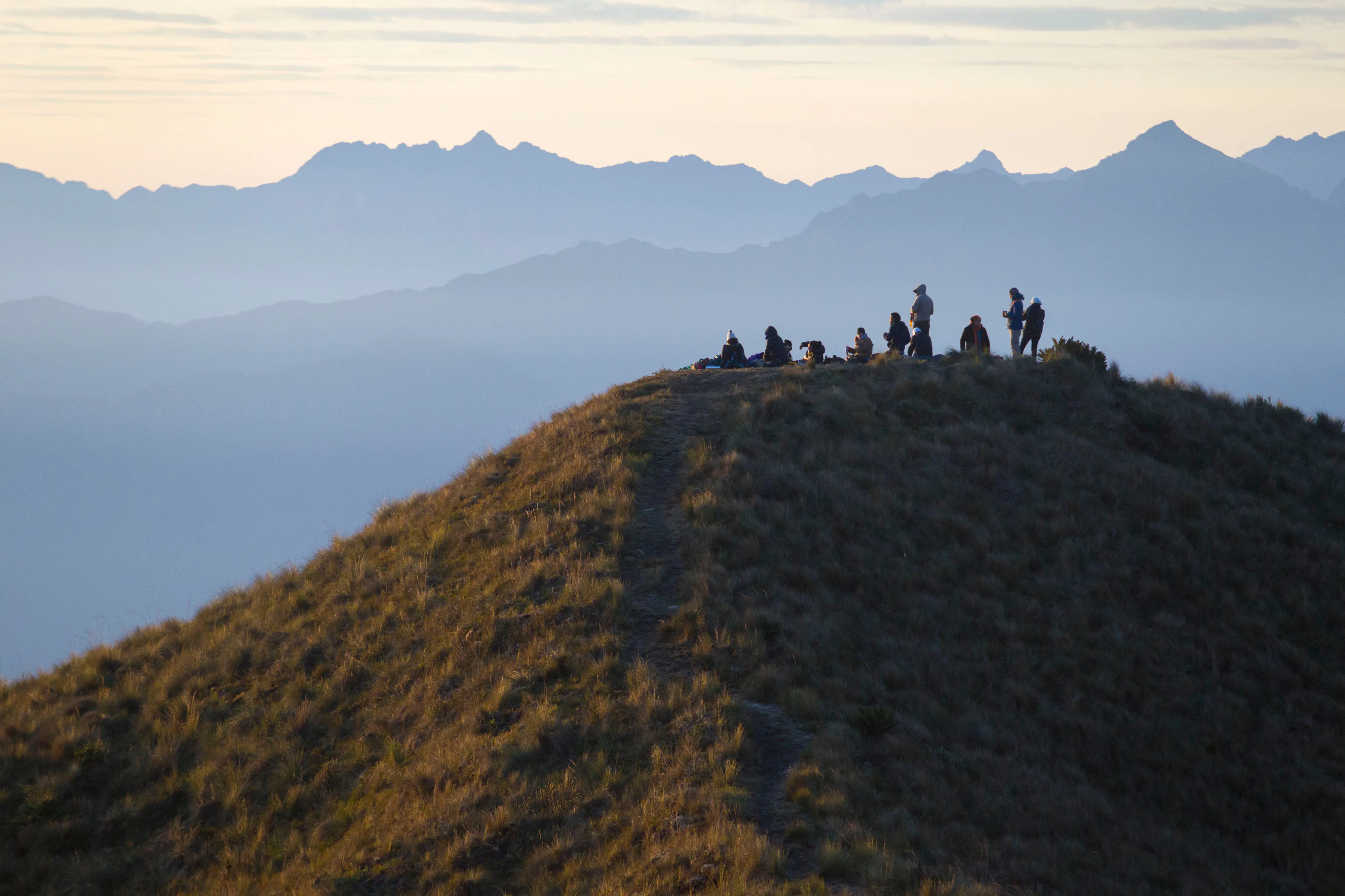 Un pico de montaña cubierto de hierba con un grupo de personas paradas encima y más montañas brumosas en la distancia.