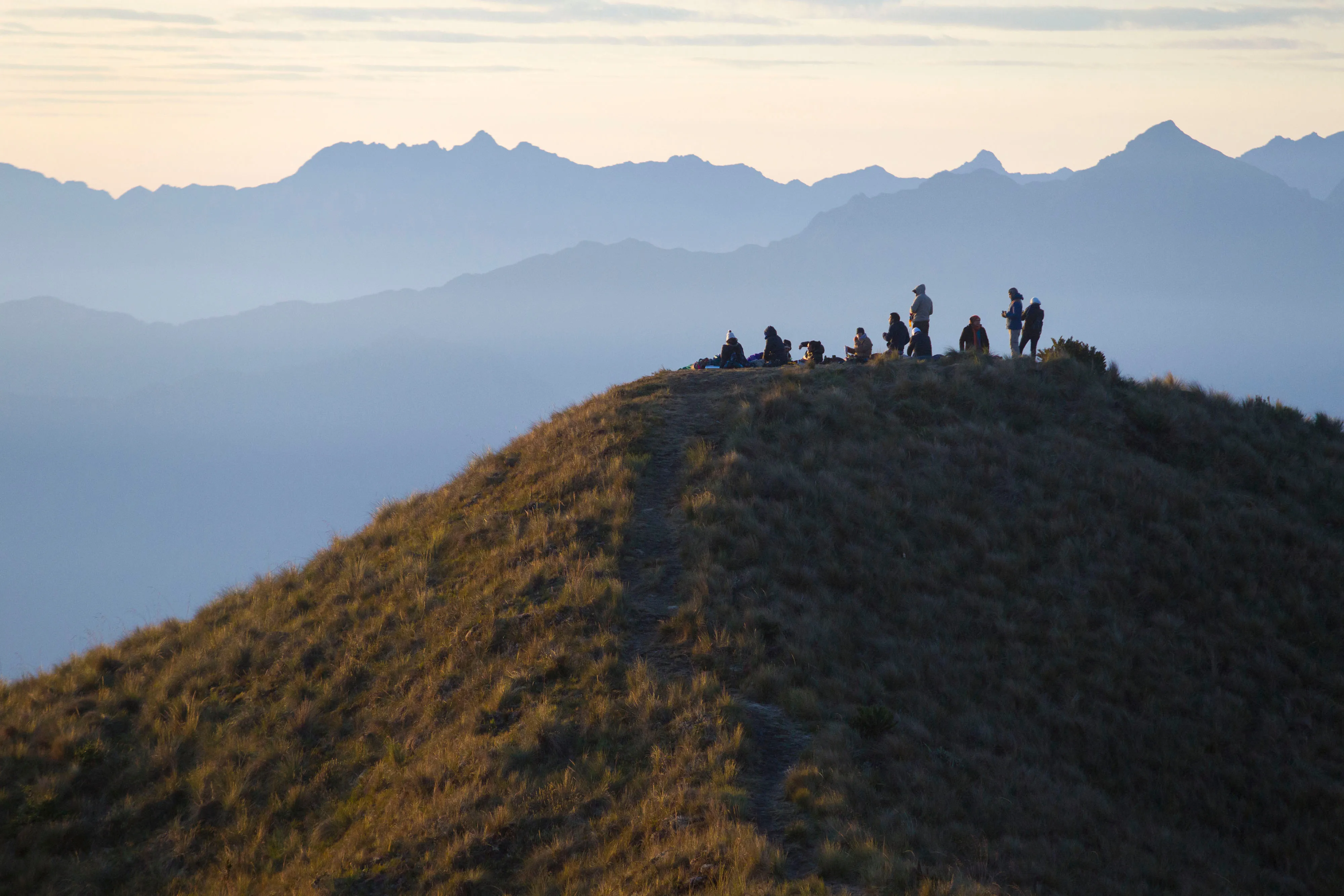 A grassy mountain peak with a group of people standing on top of it, and more hazy mountains in the distance