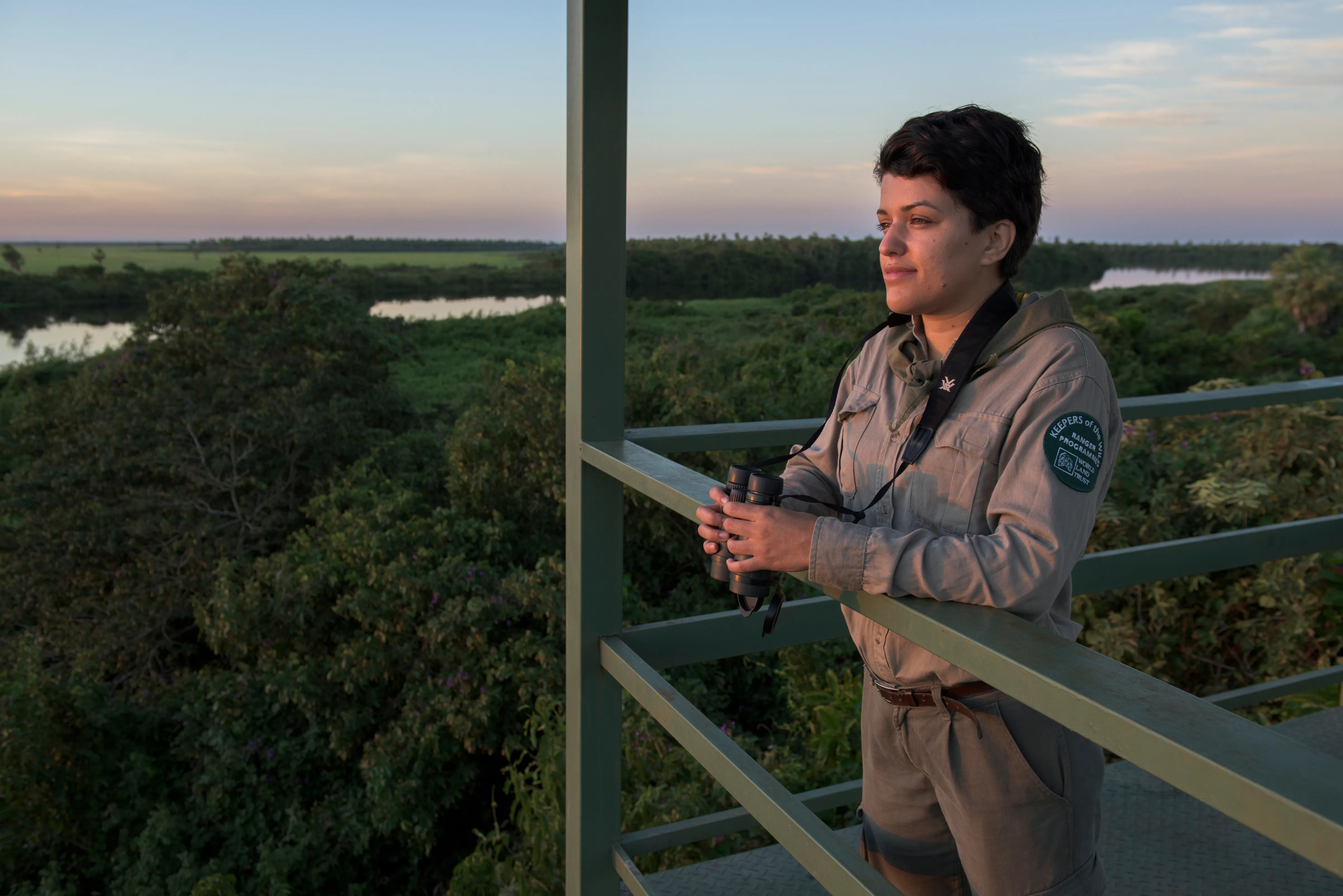 A woman holding binoculars and leaning over a tall balcony above a lush, green forest